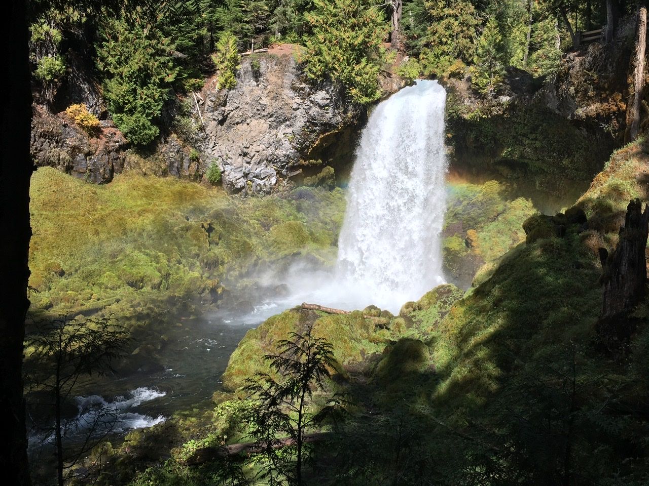 A waterfall with rainbow falling into a mossy rock-lined pool. 