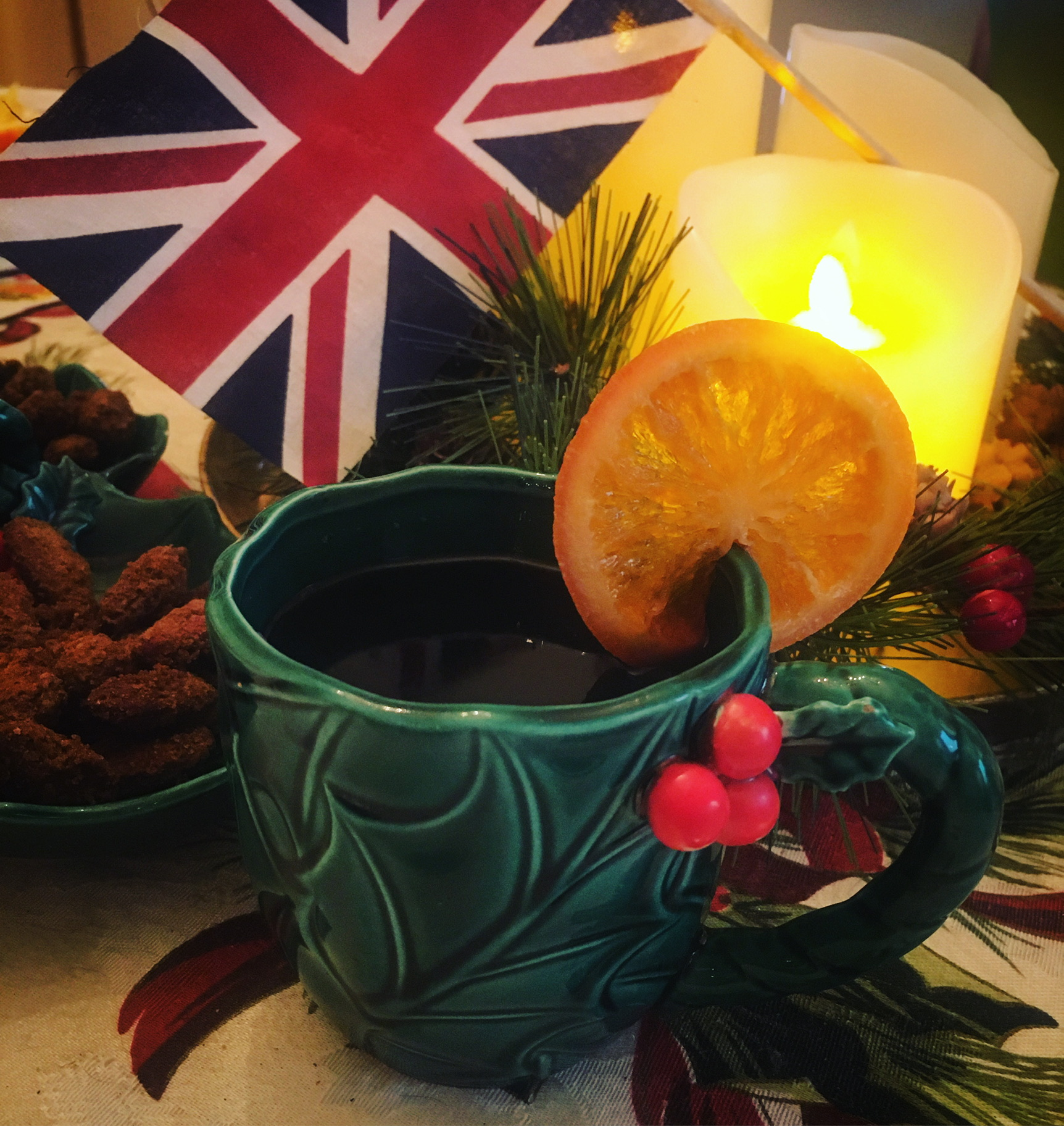 A green holly-design mug filled with a warm, wintry beverage, with the British flag, candles, and other Christmas decorations behind it.