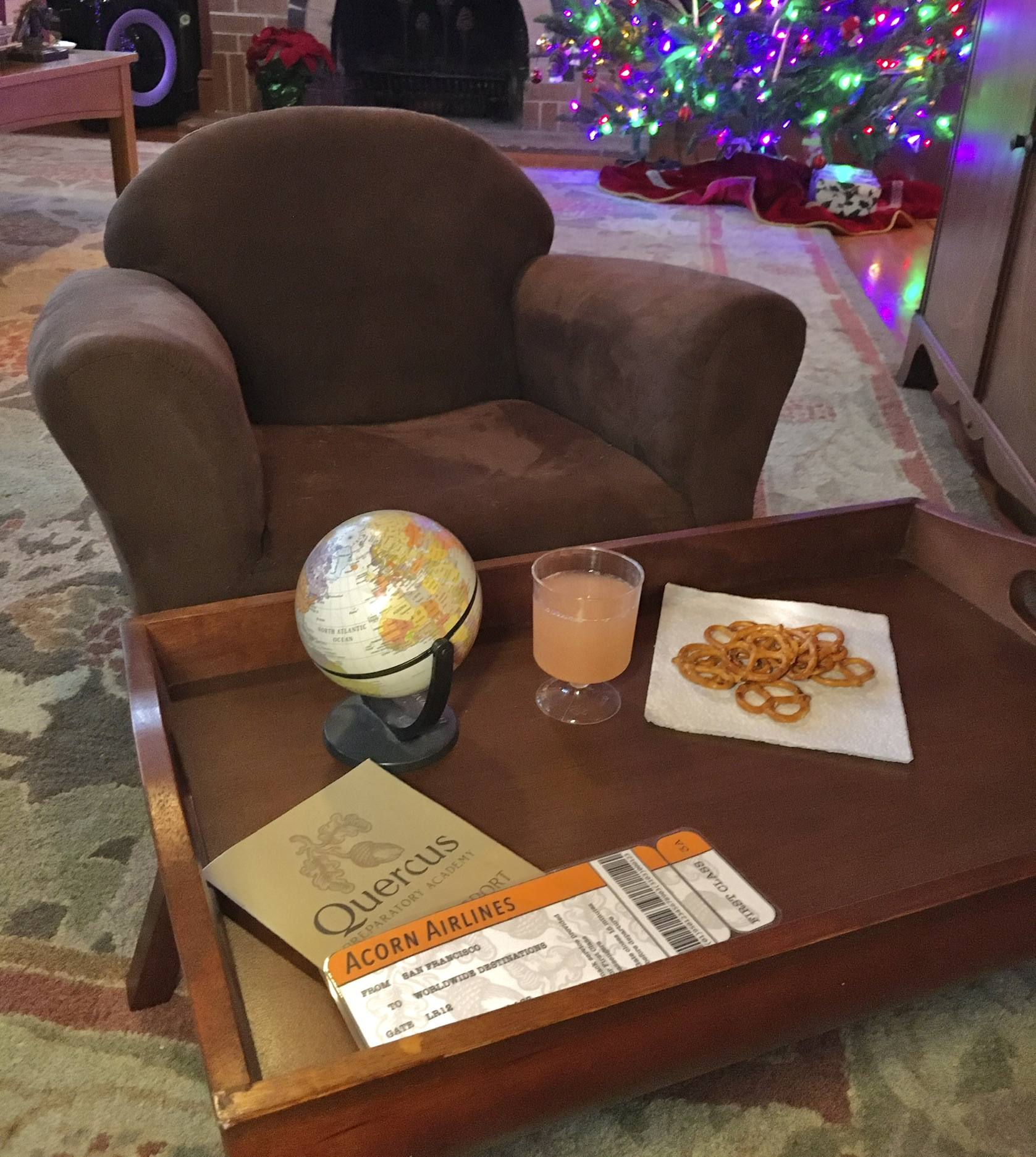 A child-size padded armchair, with a wooden tray in front holding a small drink, pretzels, a small globe, and our boarding pass and passport.
