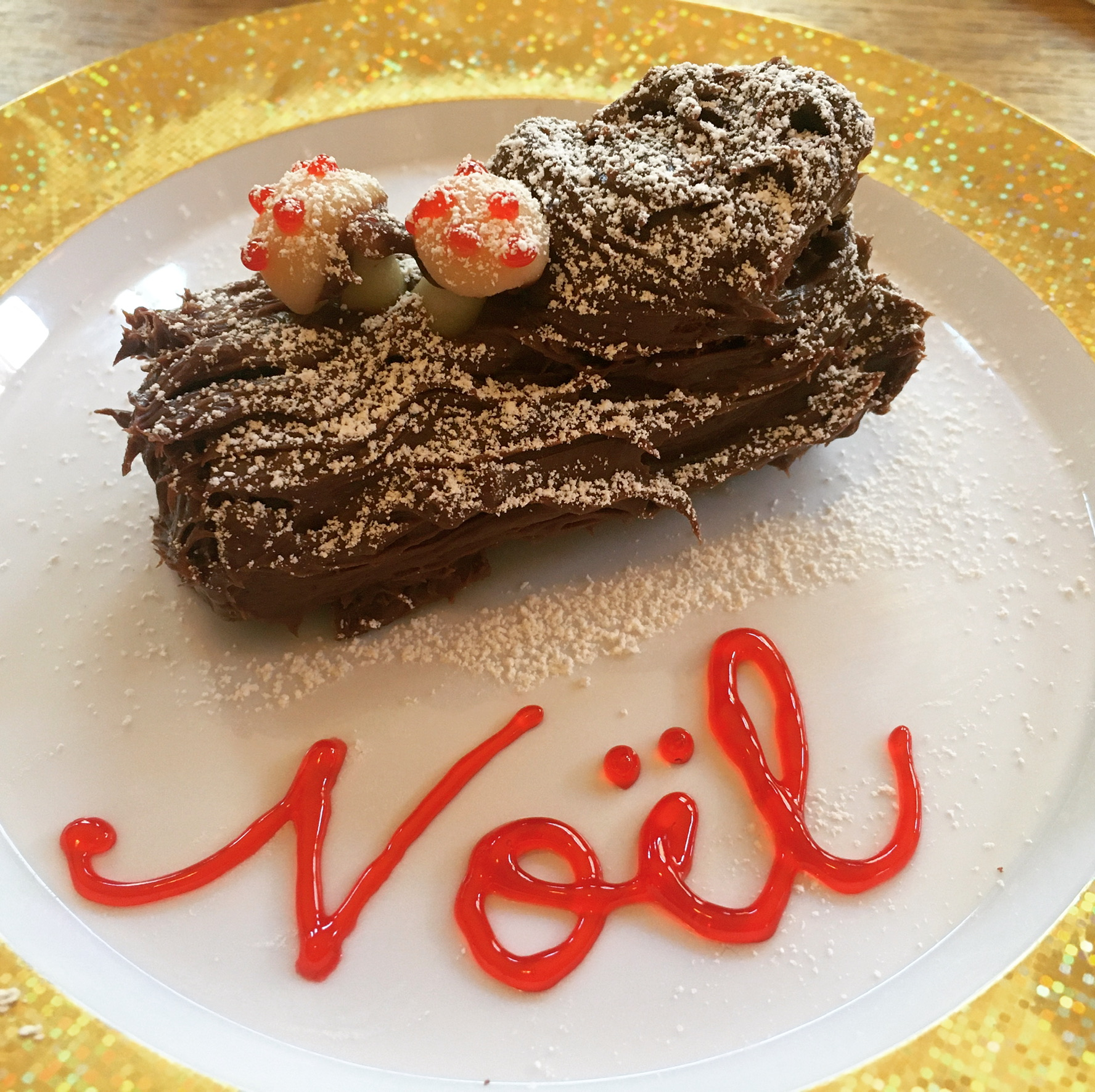 A small cake shaped like a log, dusted with powdered sugar "snow." "Noel" is written in shiny red gel.