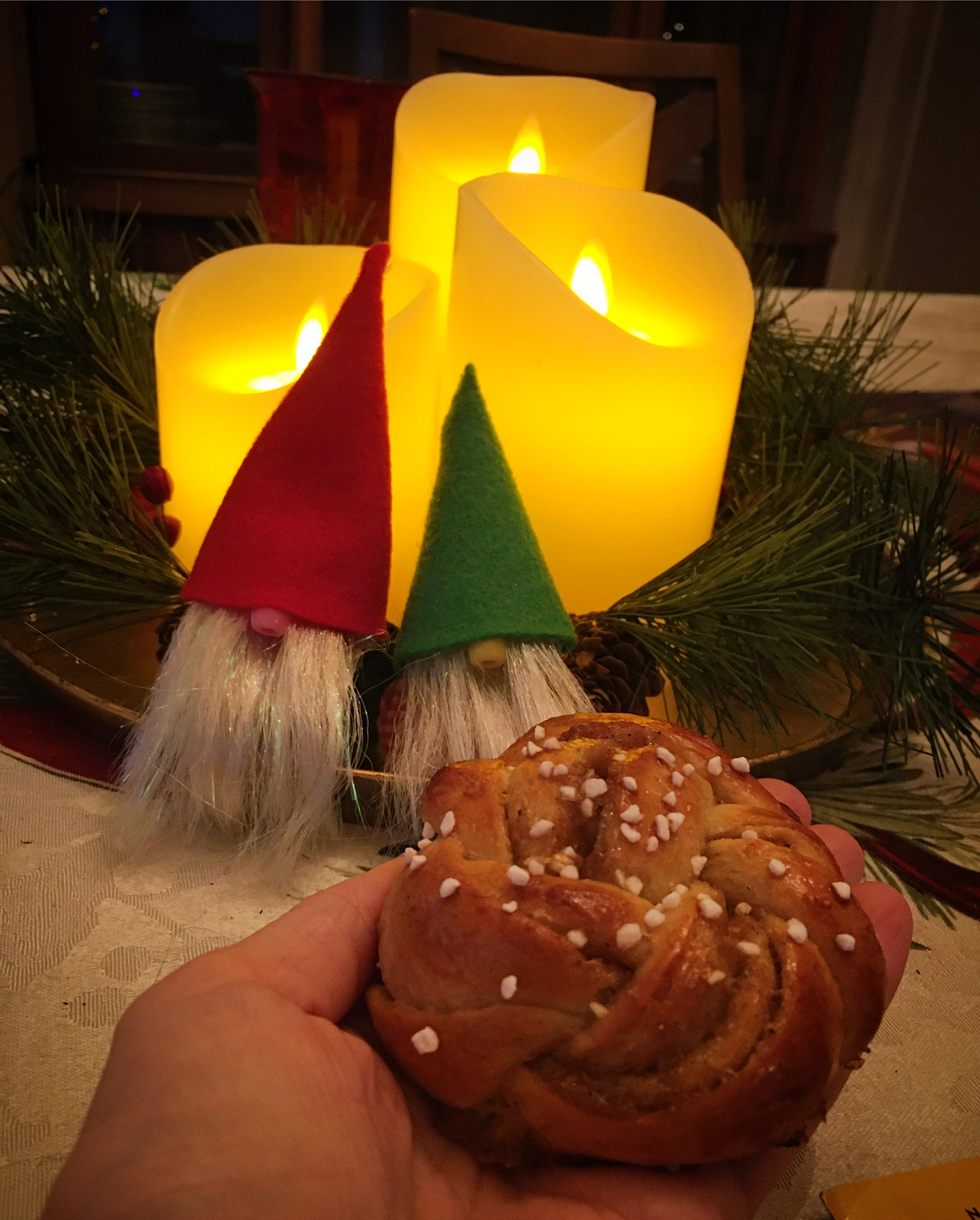 A swirled, golden brown bun is topped with white hunks of pearl sugar, and it's being offered to two little elf Nordic dolls.