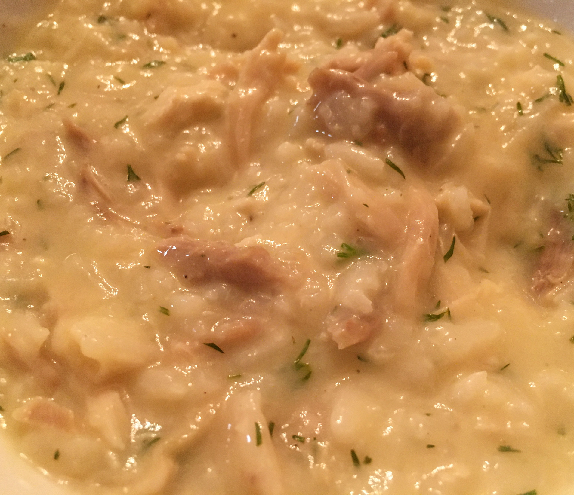 Close-up view of an opaque cream-colored soup, with bits of rice, chicken, and dill visible.