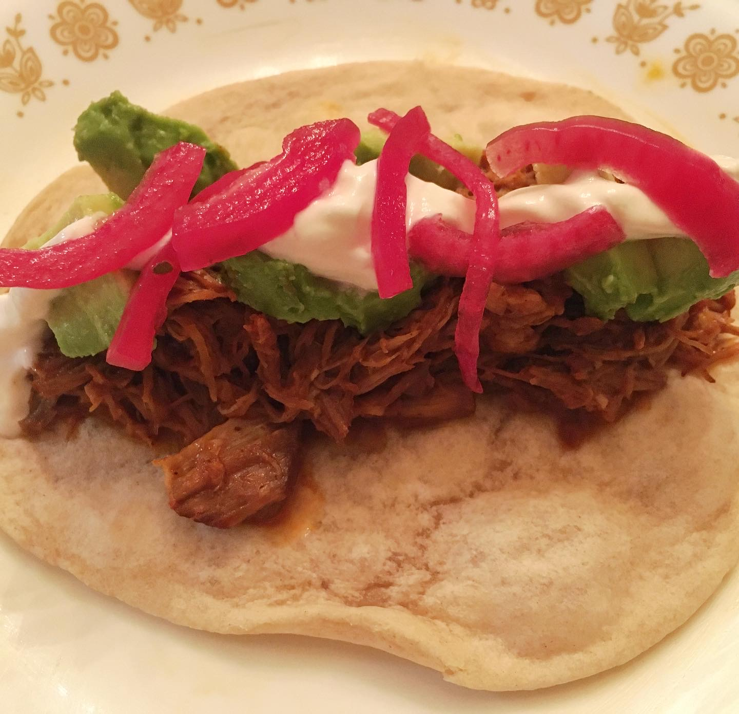 A small flour tortilla is topped with seasoned shredded pork, avocado, sour cream, and bright pink onions.