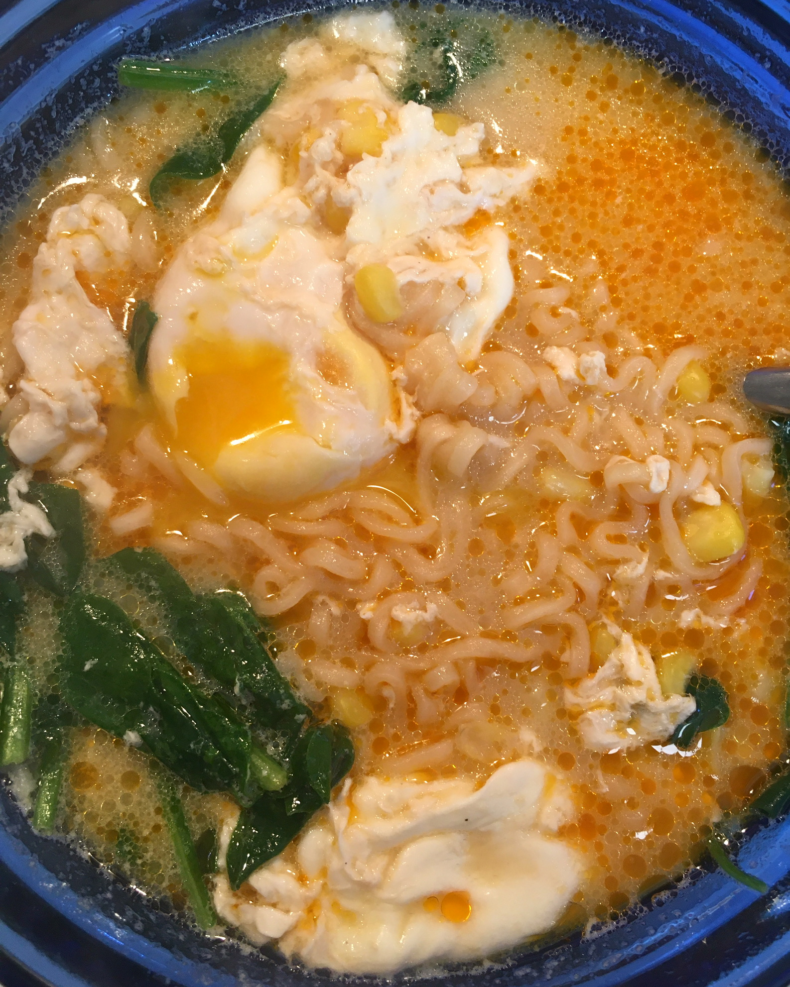 A blue bowl holds a noodle soup, with corn, poached egg, and spinach.