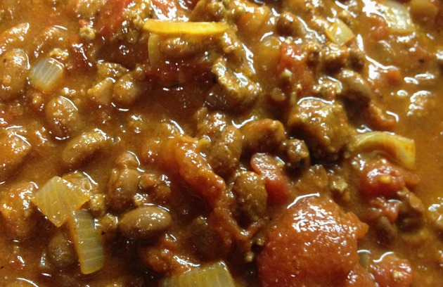 Close-up view of chili, with beans, onion, ground beef, and tomato hunks in a brown liquid.