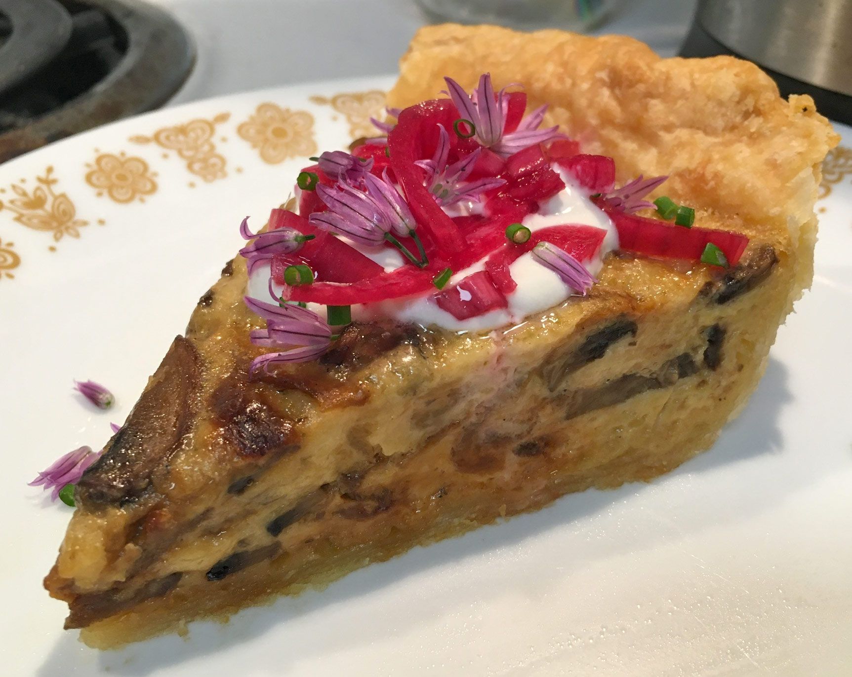 A slice of quiche is on a plate, topped with a cream sauce, bright pink onions, and small purple blossoms.