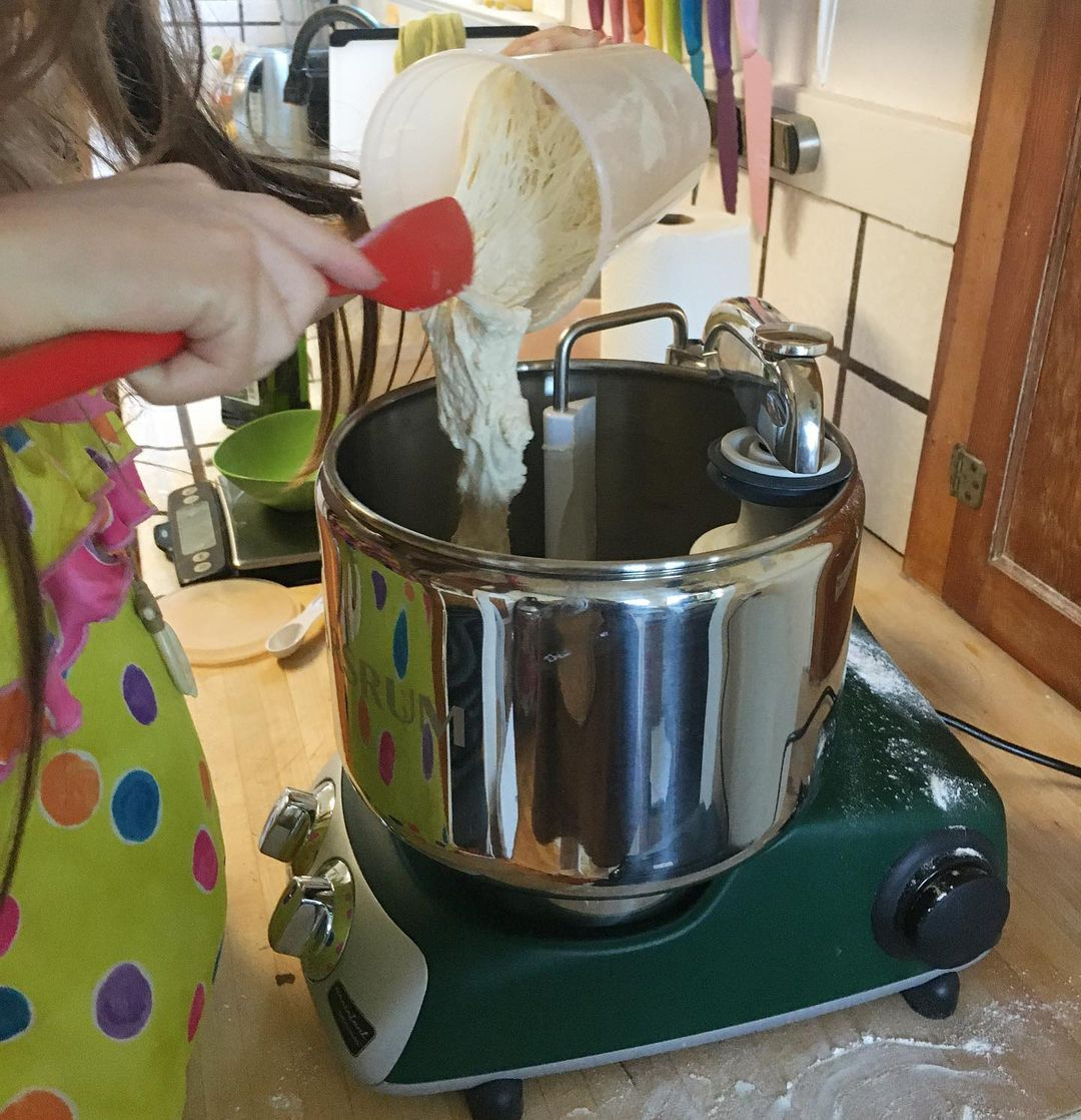 A young girl is pouring a stretchy dough into a stand mixer.