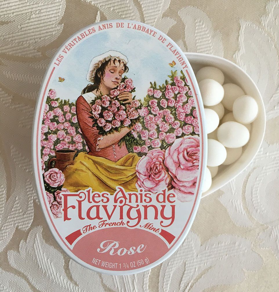 An oval tin is opened, revealing white round candies inside. The cover of the tin is a painted scene of a woman with roses.