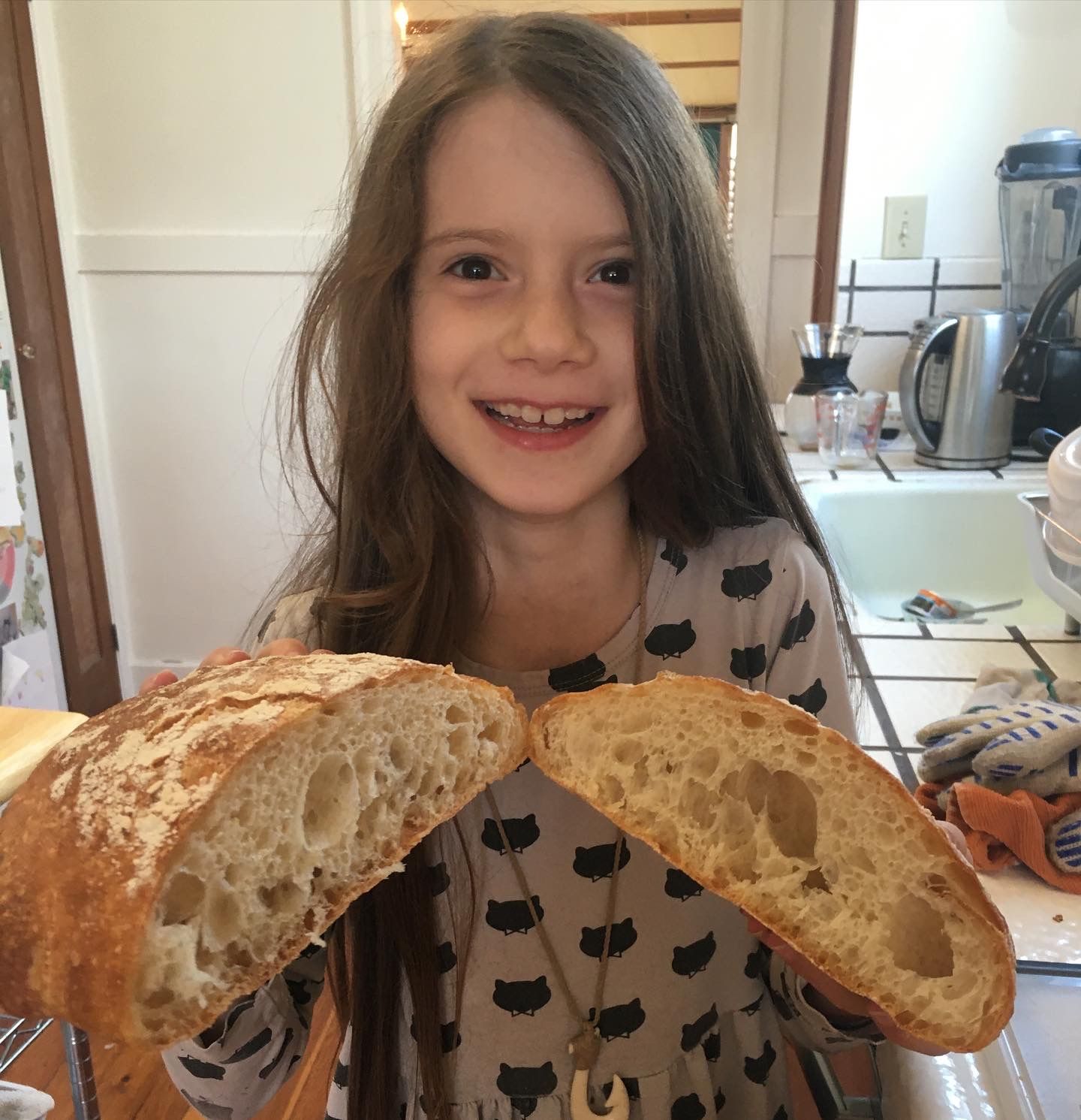 A beaming young girl is holding two halves of a sliced-open ciabatta loaf, exposing airy-textured bread inside.