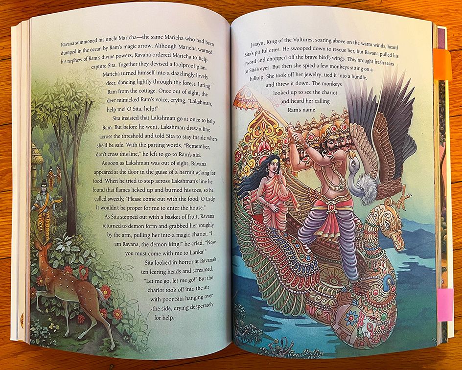 A book is open to show pages of a story from the Ramayana. There are richly detailed, colorful illustrations of Sita captive in Ravana's swan-shaped chariot, and a deer luring Ram into the forest.