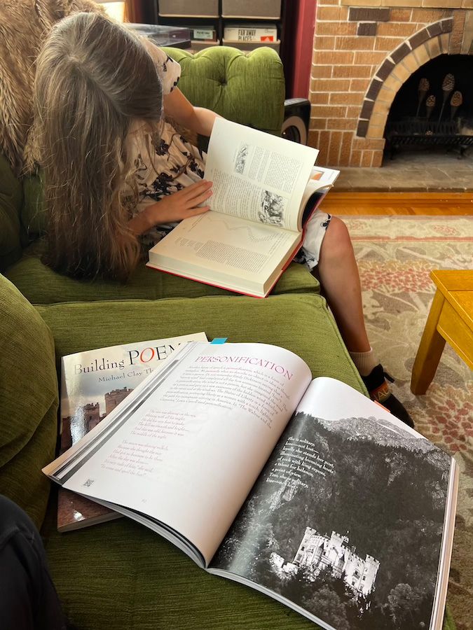 My daughter sits on a green couch reading The Annotated Alice, as our textbooks lay open in the foreground.