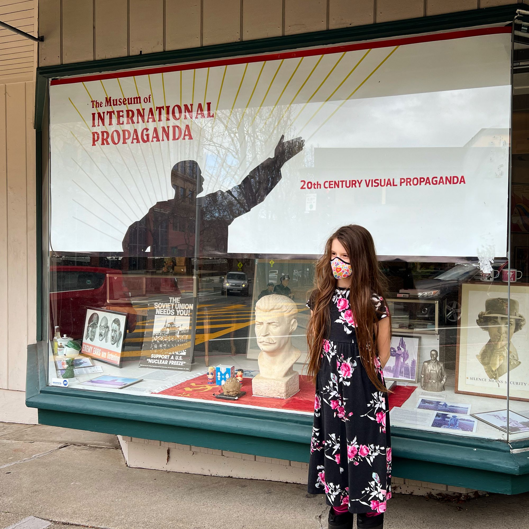 10-year-old Wanda, wearing a long black dress with pink roses, and with very long hair, stands in front of the main window to the Museum of International Propaganda.