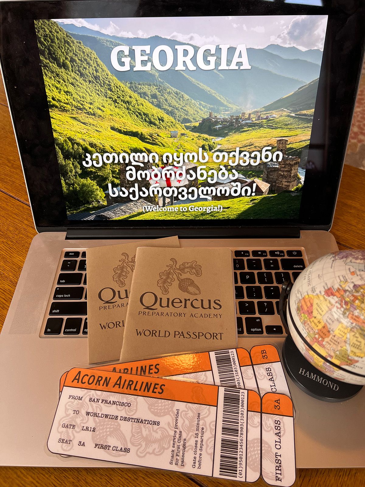 Laptop screen showing a presentation saying "Welcome to Georgia!," with a Quercus Prep passport and Acorn Airlines plane tickets on the keyboard.