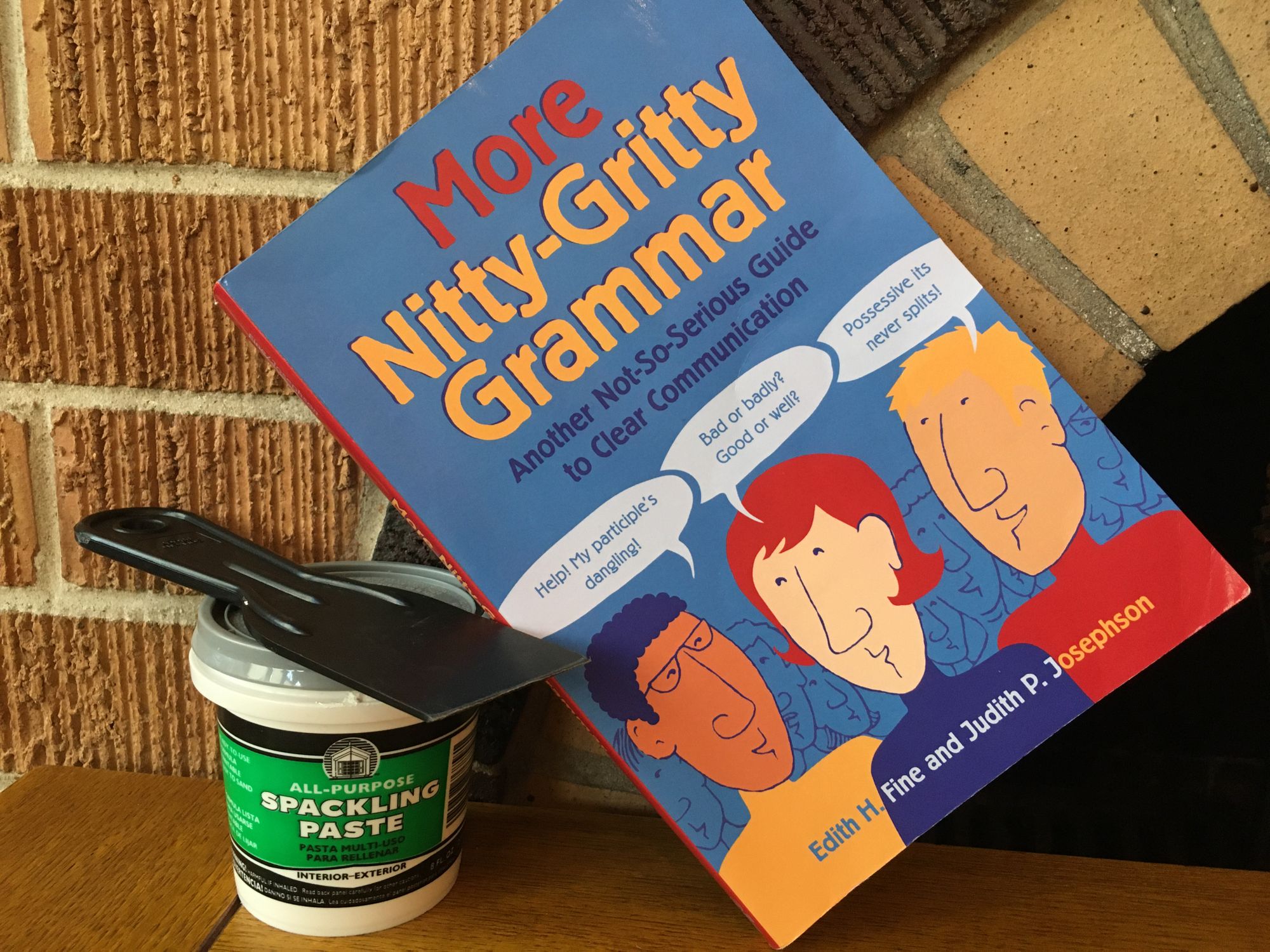 A paperback book titled Nitty Gritty Grammar is balanced on a small container of spackling paste, along with a putty knife.