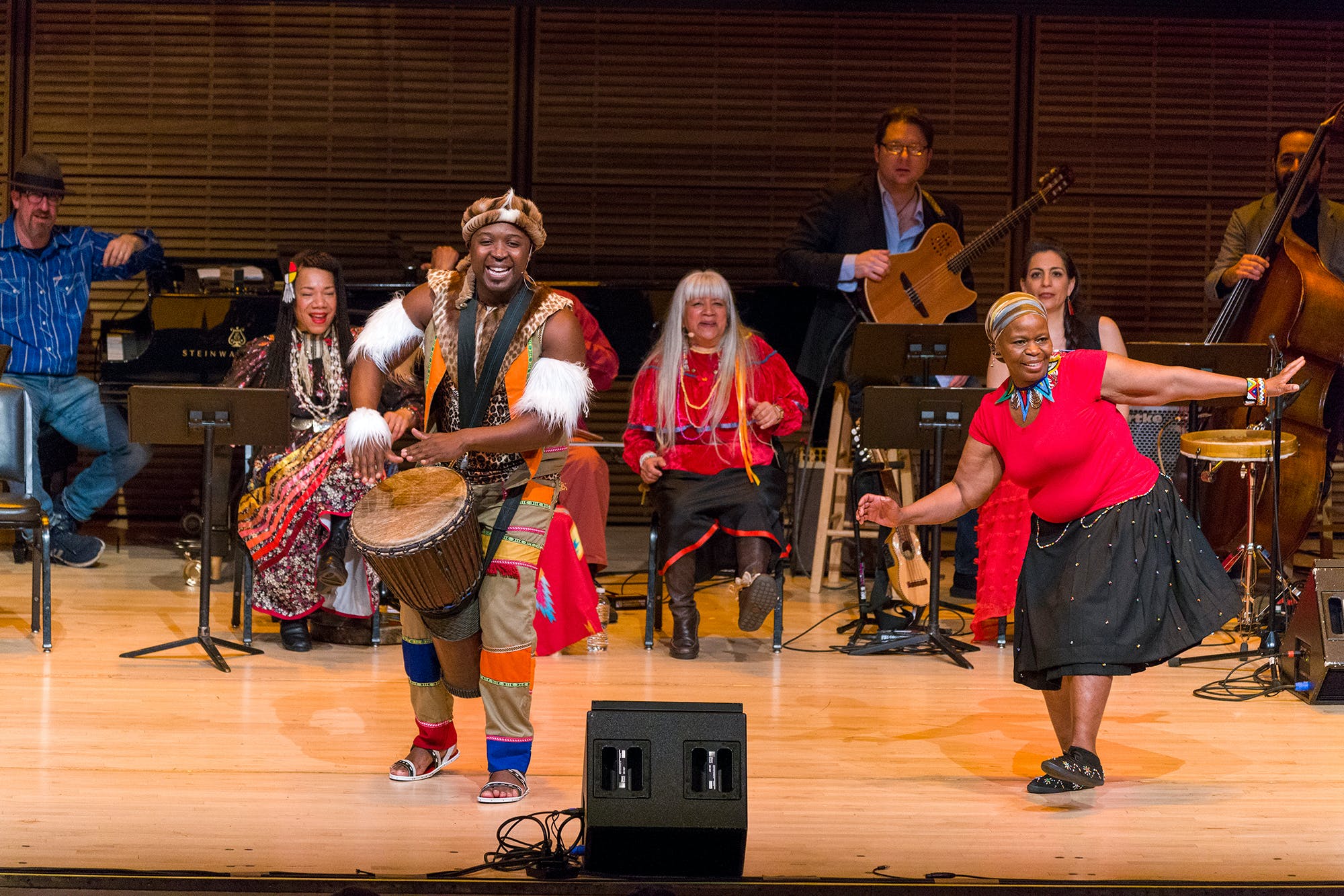 Musicians performing on stage. Some are wearing Native American ribbon dresses, some are wearing African Zulu garb.